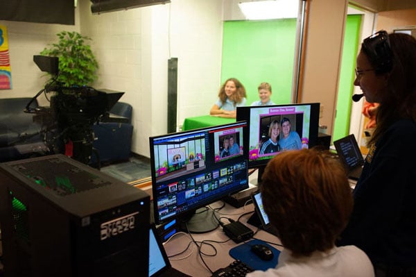 A media center with a computer display in the foreground and two students sitting in front of a green screen.