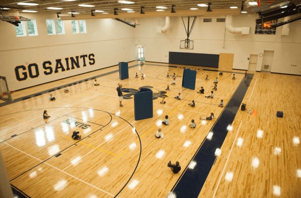 A gym with students sitting down, doing a physical activity with a "Go Saints" decal on the wall.